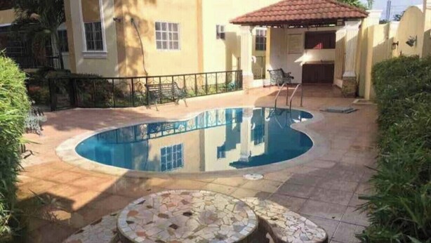 Ein Schwimmbecken mit Fliesen umgeben, ein Tisch mit Mosaikdekor und im Hintergrund ein Gebäude mit roten Ziegeldach.
