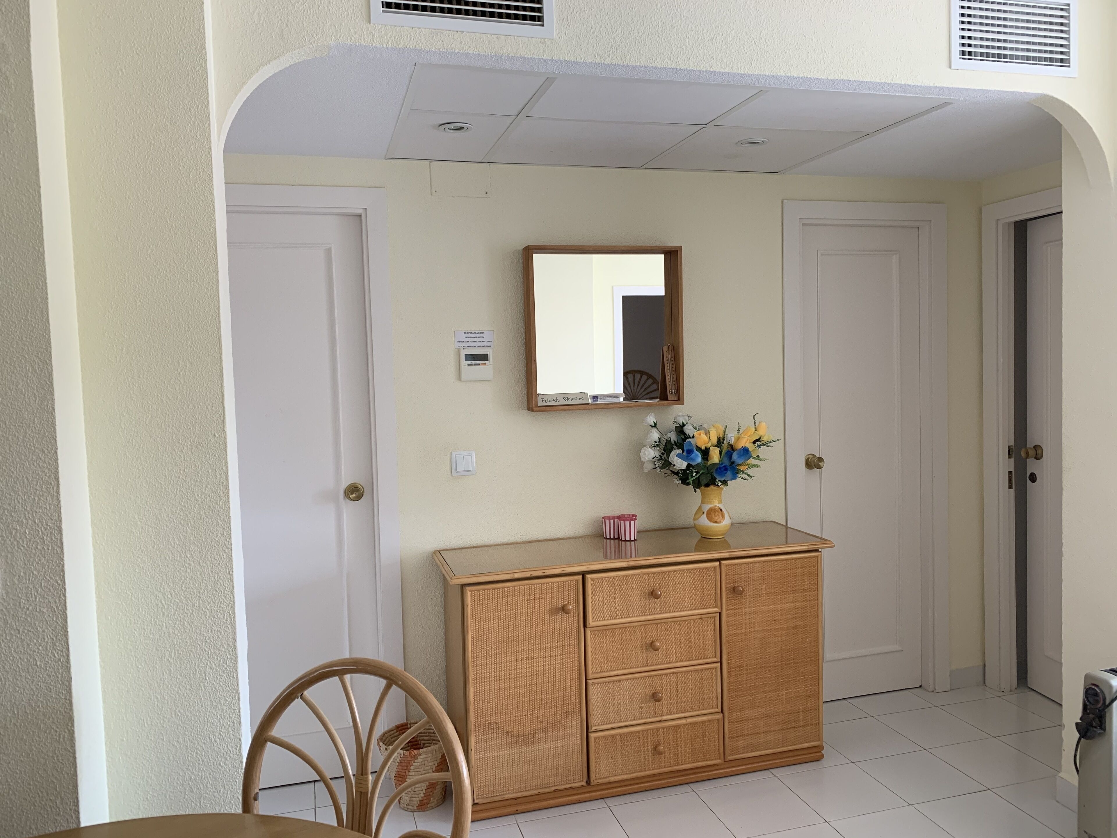 A room with a rattan cabinet, a mirror, a vase with flowers, a chair, and a door.