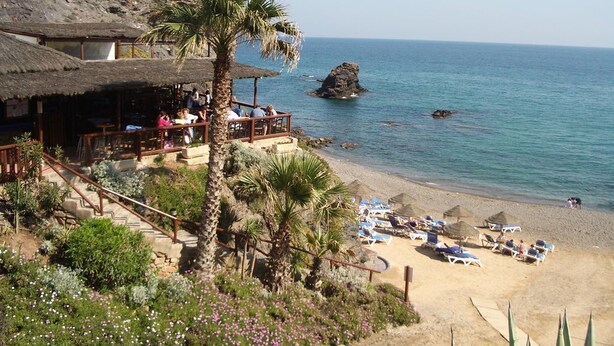 Una località balneare con un edificio a tetto di paglia, palme e un promontorio roccioso nel mare.