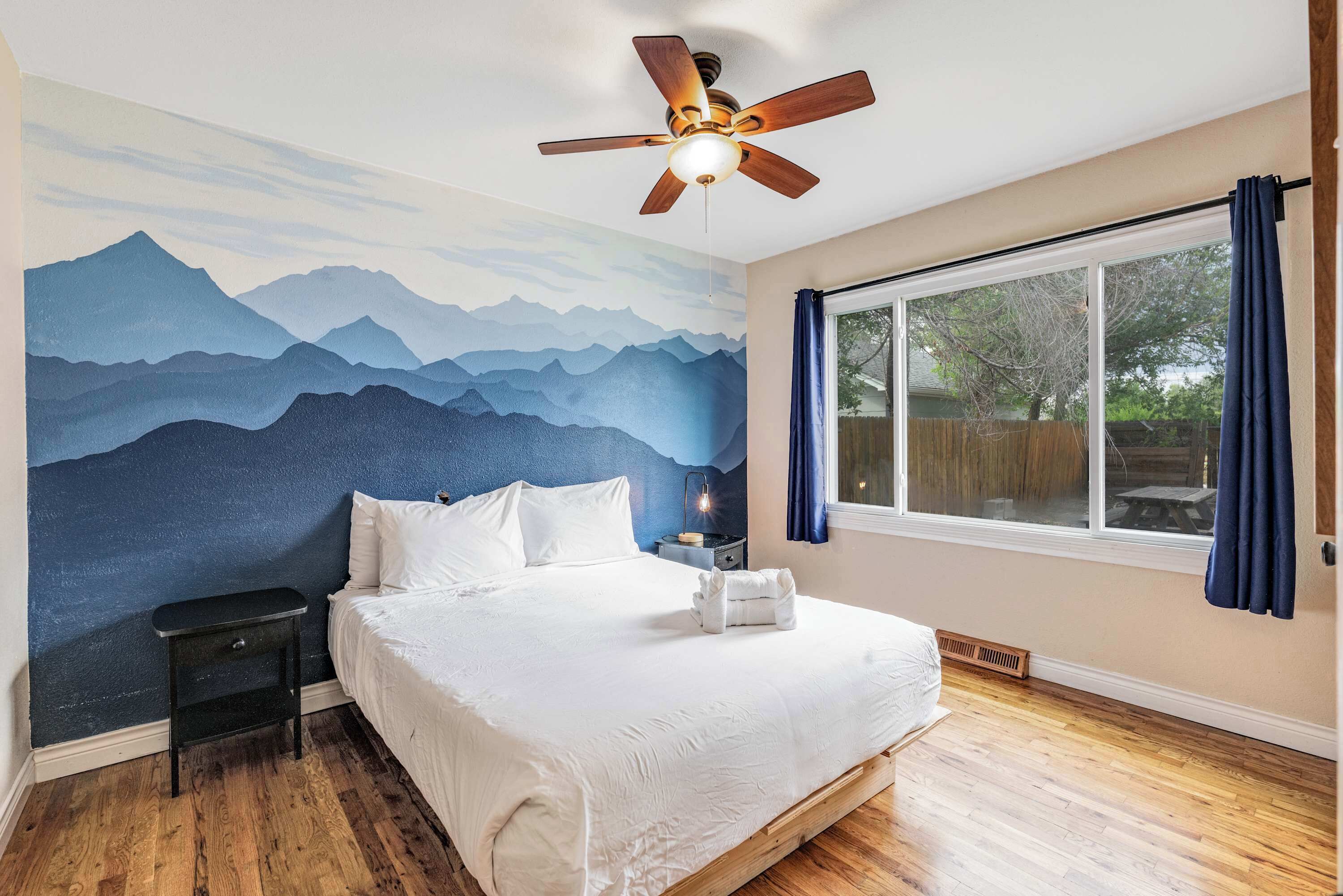 Una camera da letto con un murale raffigurante montagne, una ventola a soffitto, un letto con lenzuola bianche, un comodino e un'ampia finestra con tende.