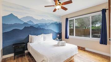 A bedroom with a mountain mural, a ceiling fan, a bed with white linens, a nightstand, and a large window with curtains.