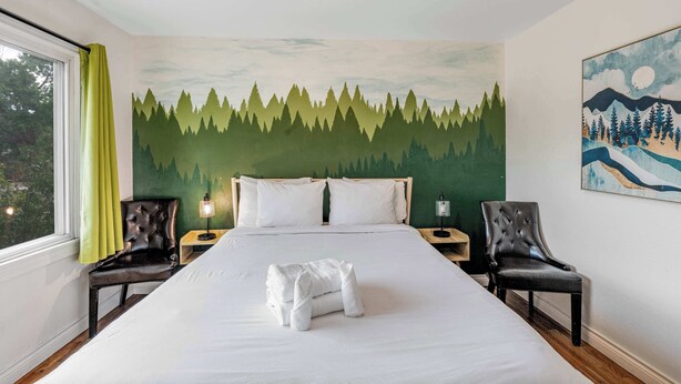 A hotel room with a bed, two chairs, a nightstand with a lamp, a window with curtains, and a forest-themed wall mural. - 2BR Pet friendly BBQ Fire Pit in EastSide Colorado (Colorado Springs)