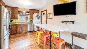 A modern kitchen with wooden cabinets, a stainless steel refrigerator, a washing machine, and a dining area with a white table and colorful stools. - 2BR Pet friendly BBQ Fire Pit in EastSide Colorado (Colorado Springs)