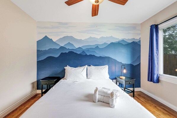A bedroom with a mountain mural, a bed with white linens, a wooden ceiling fan, a bedside table with a lamp, and a window with blue curtains.