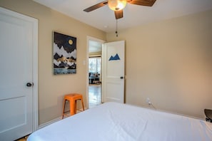 Un dormitorio con cama, ventilador de techo, un cuadro y una puerta que da a otra habitación.