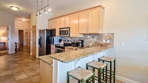 A kitchen with wooden cabinets, granite countertops, a refrigerator, a microwave, a stove, and a sink. There are three bar stools and a chandelier.