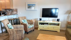 A living room with wicker chairs, a television, a wooden coffee table, and a small kitchen area with a sink and cabinets.