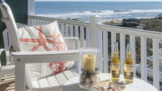 A white deck chair with a starfish pillow, two bottles of Corona Extra beer, and a candle in a glass jar on a table overlooking the ocean.