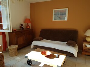 Una sala de estar acogedora con un sofá marrón, una mesita con un control remoto, un taburete de mimbre y un cuadro en la pared.