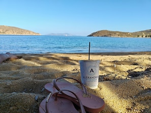 Escena de playa con un vaso de bebida con la etiqueta "ALEXANDROS BY THE BEACH SERIFOS ISL" y chanclas en la arena.