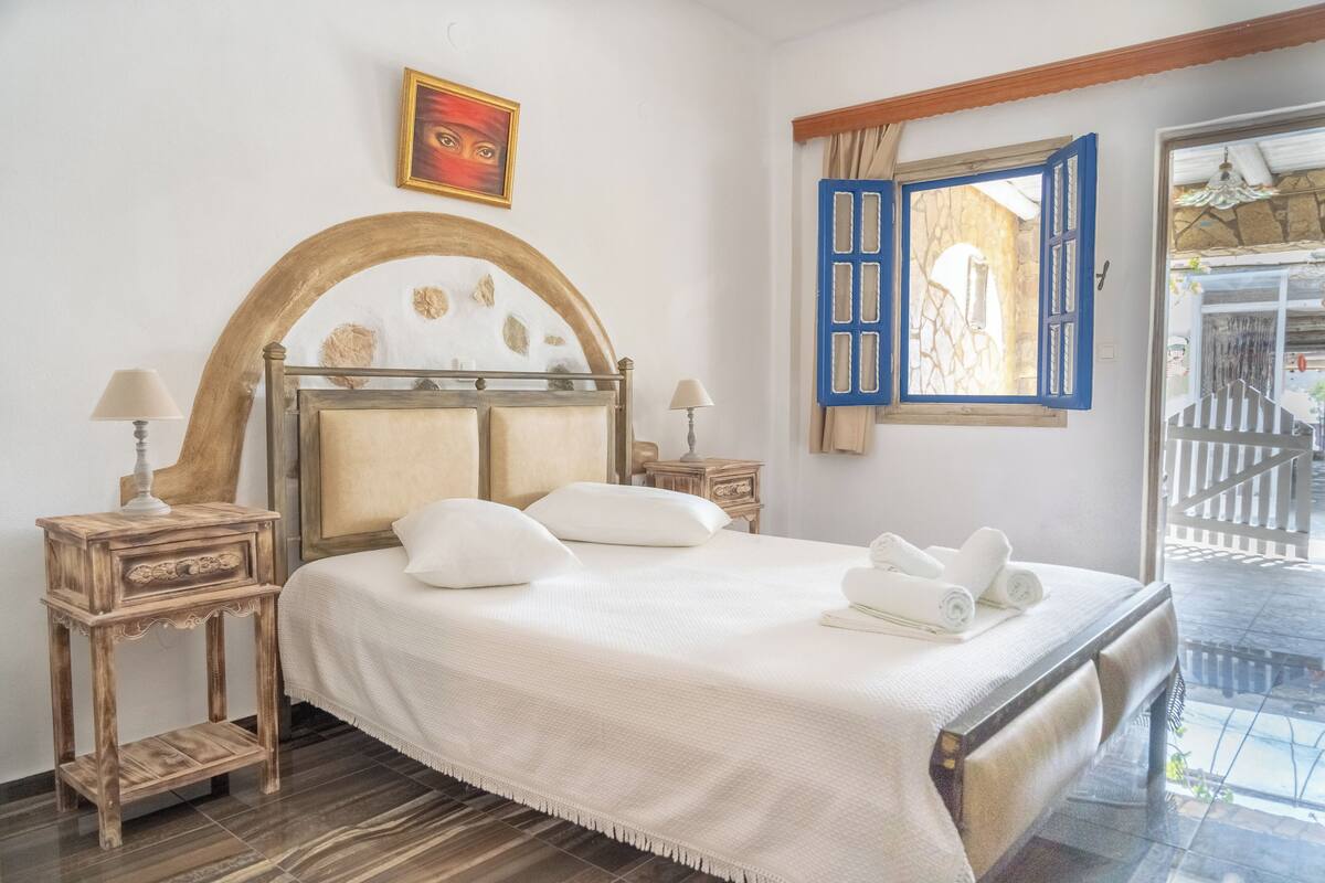 A bedroom with a large bed, two bedside tables, a painting, a window with blue shutters, and a view of the outside.