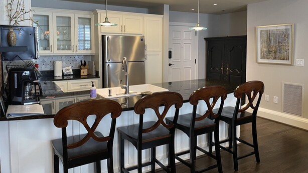 Cocina moderna con armarios blancos, refrigerador de acero inoxidable, una isla central con taburetes altos, iluminación colgante y un amplio ventanal.