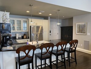 A modern kitchen with white cabinets, a stainless steel refrigerator, a central island with bar stools, pendant lighting, and a large window.