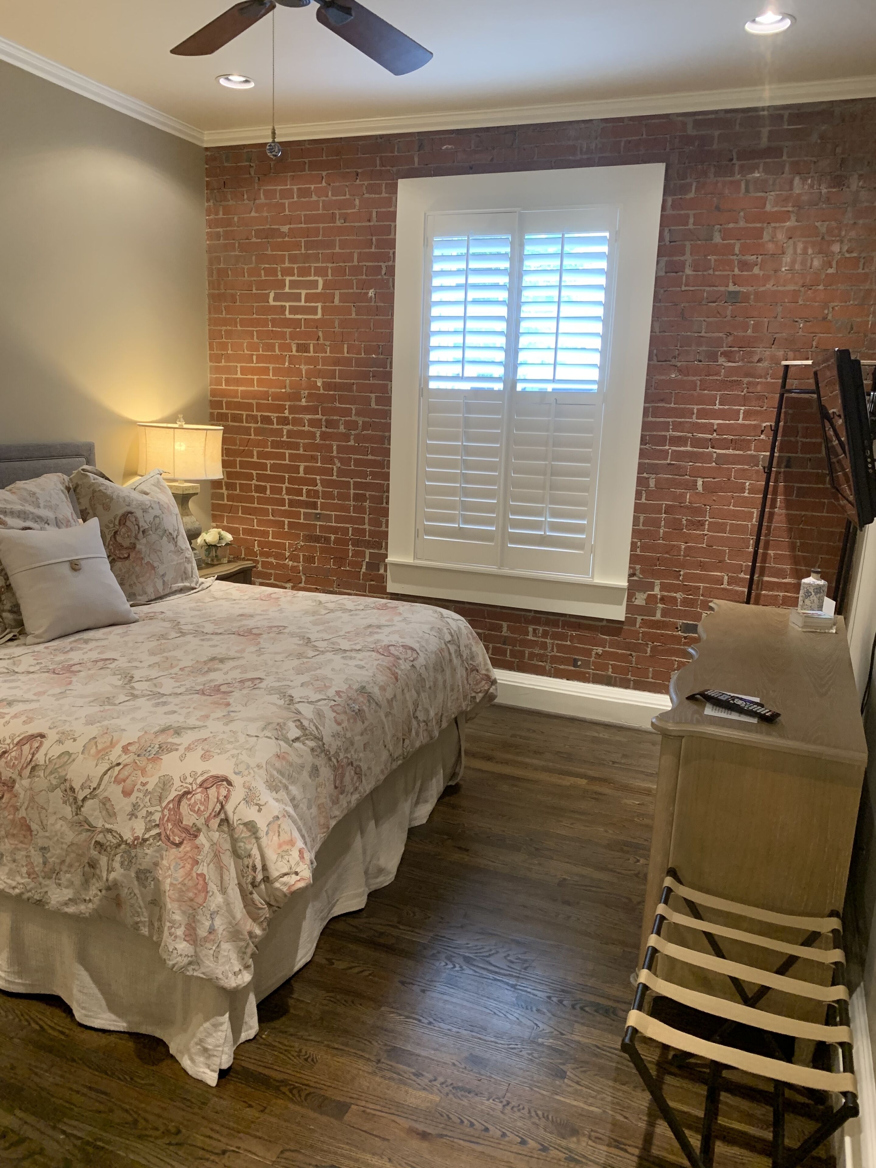 A bedroom with a brick wall, a bed with floral bedding, a nightstand with a lamp, and a wooden dresser.