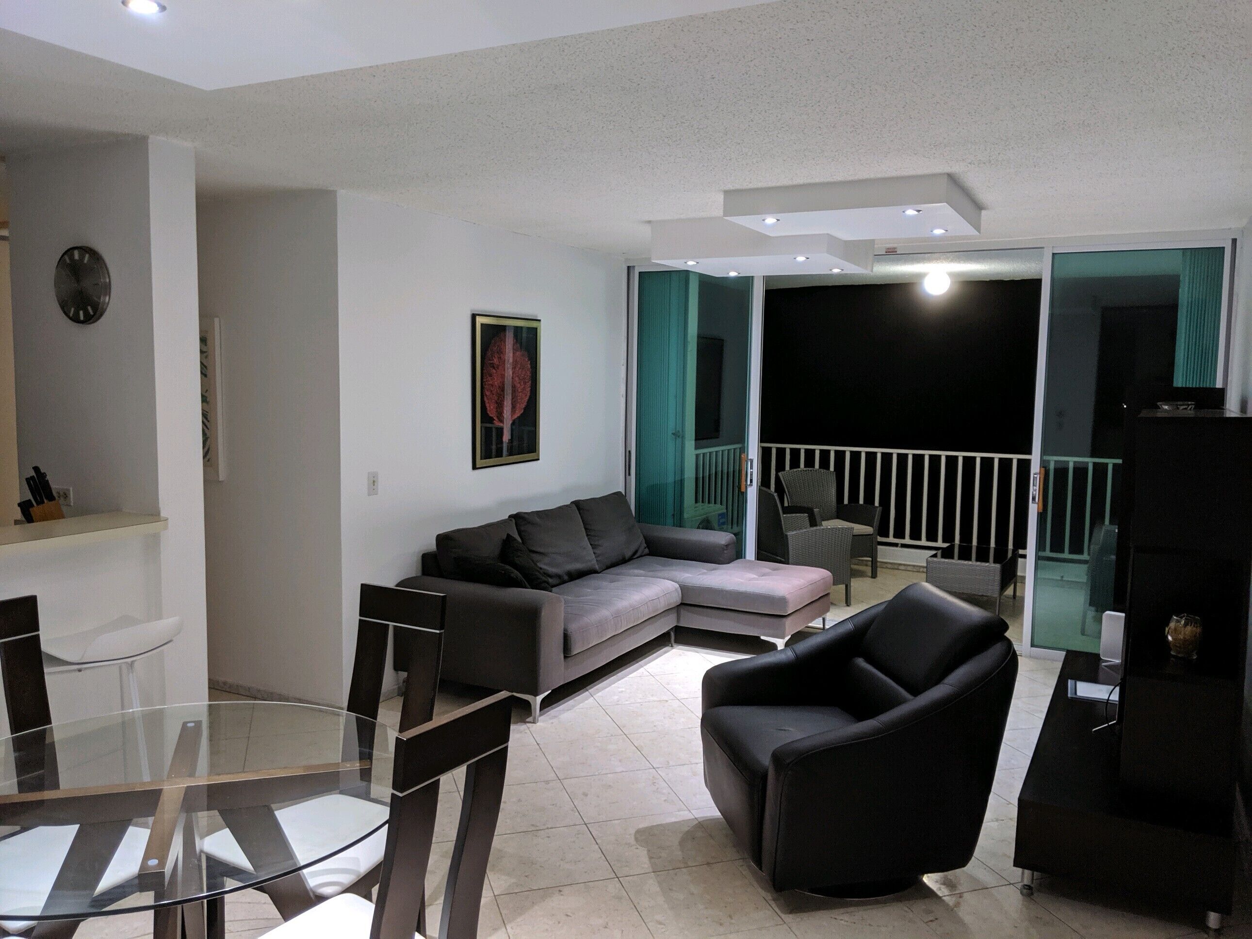 Una sala moderna con una mesa de comedor de vidrio, un sofá, un sillón negro y un televisor sobre un mueble.
