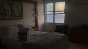 Ein schlafwagenzimmer in gedämpftem Licht, mit Bett, Sessel, Kommode und Fenster mit Meerblick.