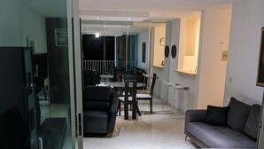 Ein modernes Wohnzimmer mit Glastrennwand, einer Couch, einem Essbereich mit Tisch und Stühlen sowie einer Küchenzeile mit Theke und Hockern.