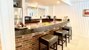 A kitchen with a brick backsplash, a stainless steel refrigerator, a microwave, and a sink. There are three bar stools and a bar counter.