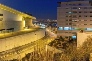 Un bâtiment moderne à la façade incurvée, doté d’un espace de stationnement et offrant une vue sur le paysage urbain lointain, de nuit.