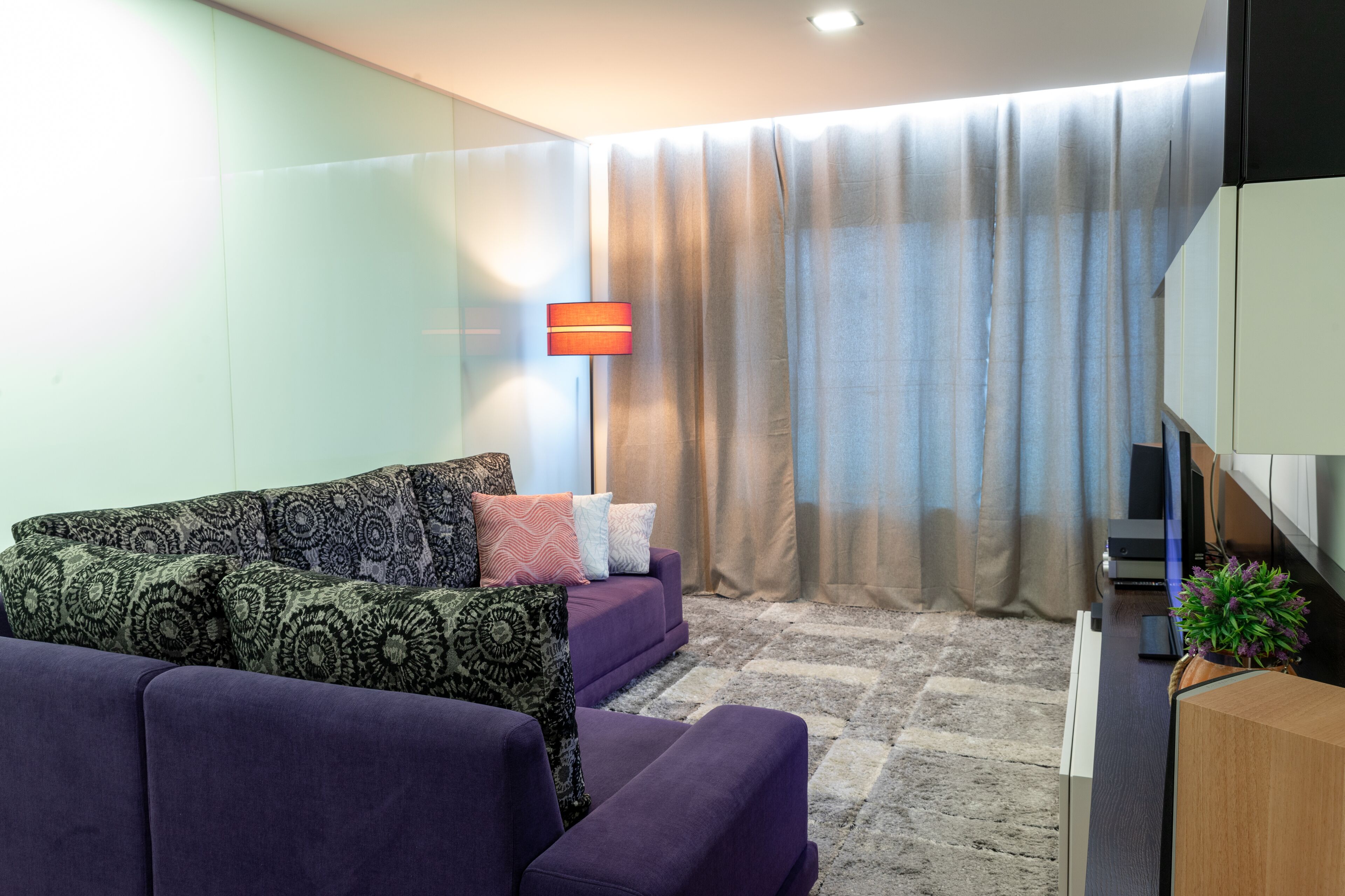 Una sala de estar moderna con un sofá morado, cojines estampados, un televisor de pantalla plana y un ventanal con cortinas.