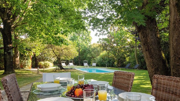 A table set for breakfast with plates, glasses, and a fruit bowl, overlooking a swimming pool and garden chairs. - Ancient Villa Art Nouveau original, large park and pool at 3 Km from Lucca Walls (Capannori)