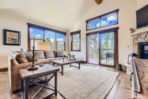 A spacious living room with a sofa, coffee table, and fireplace, large windows, and a stone fireplace surround.