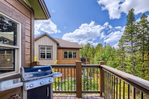 A wooden deck with a grill, a house in the background, and a forested area.