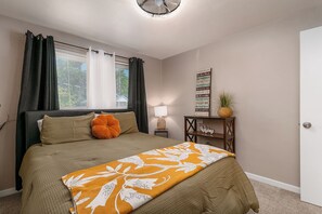 Een slaapkamer met een bed, gordijnen, een bedlamp, een welkomstbord en een klein tafeltje met een plant.