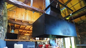 A rustic interior with a large black stove, wooden beams, stone walls, and a cozy seating area with cushions.