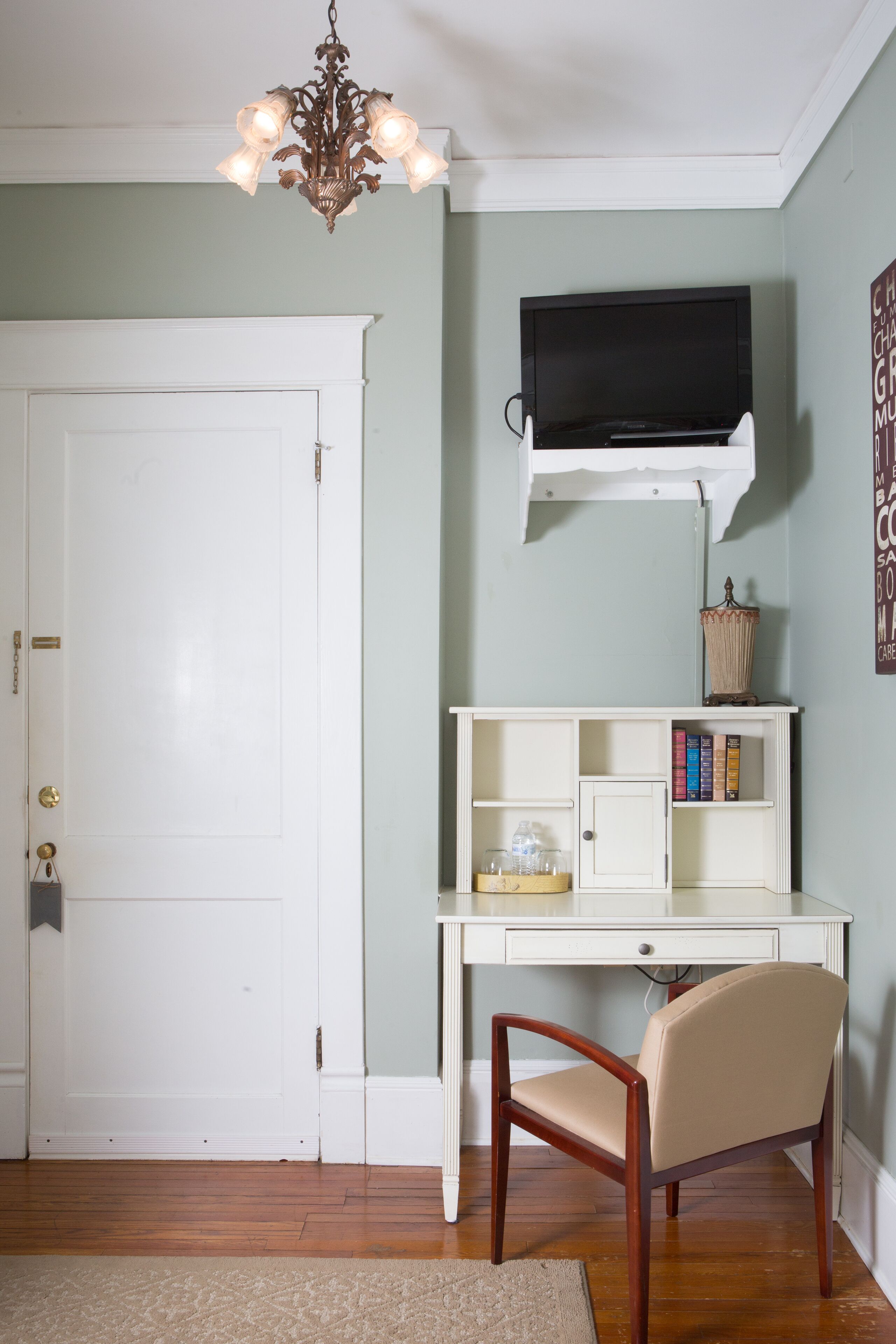 A room with a white door, a desk with a chair, a mounted TV, and a small shelf with books and decorative items.