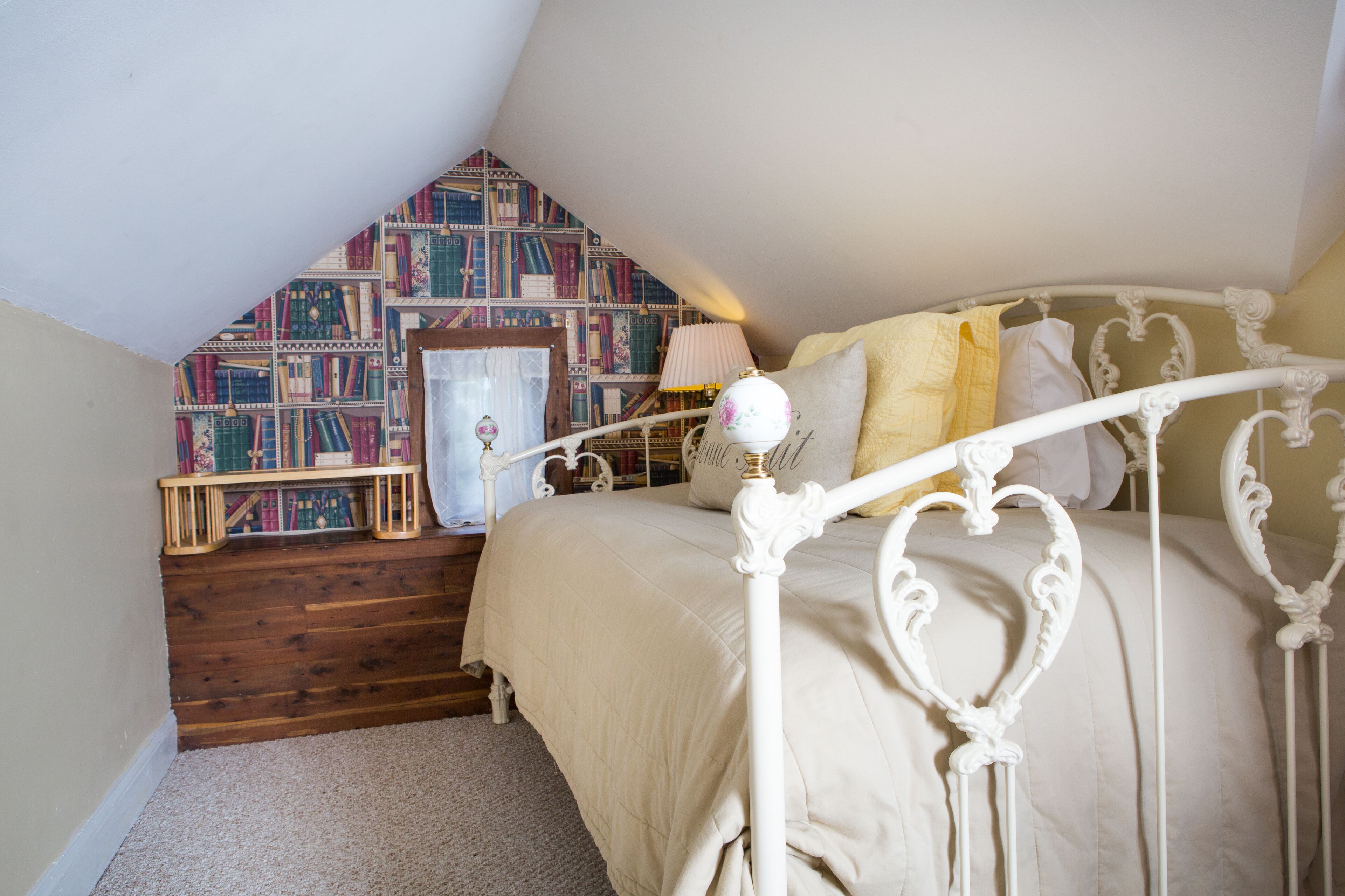 Un dormitorio con techo inclinado, estructura de cama de metal blanco, cubrecama color beige y estantería con diseño.