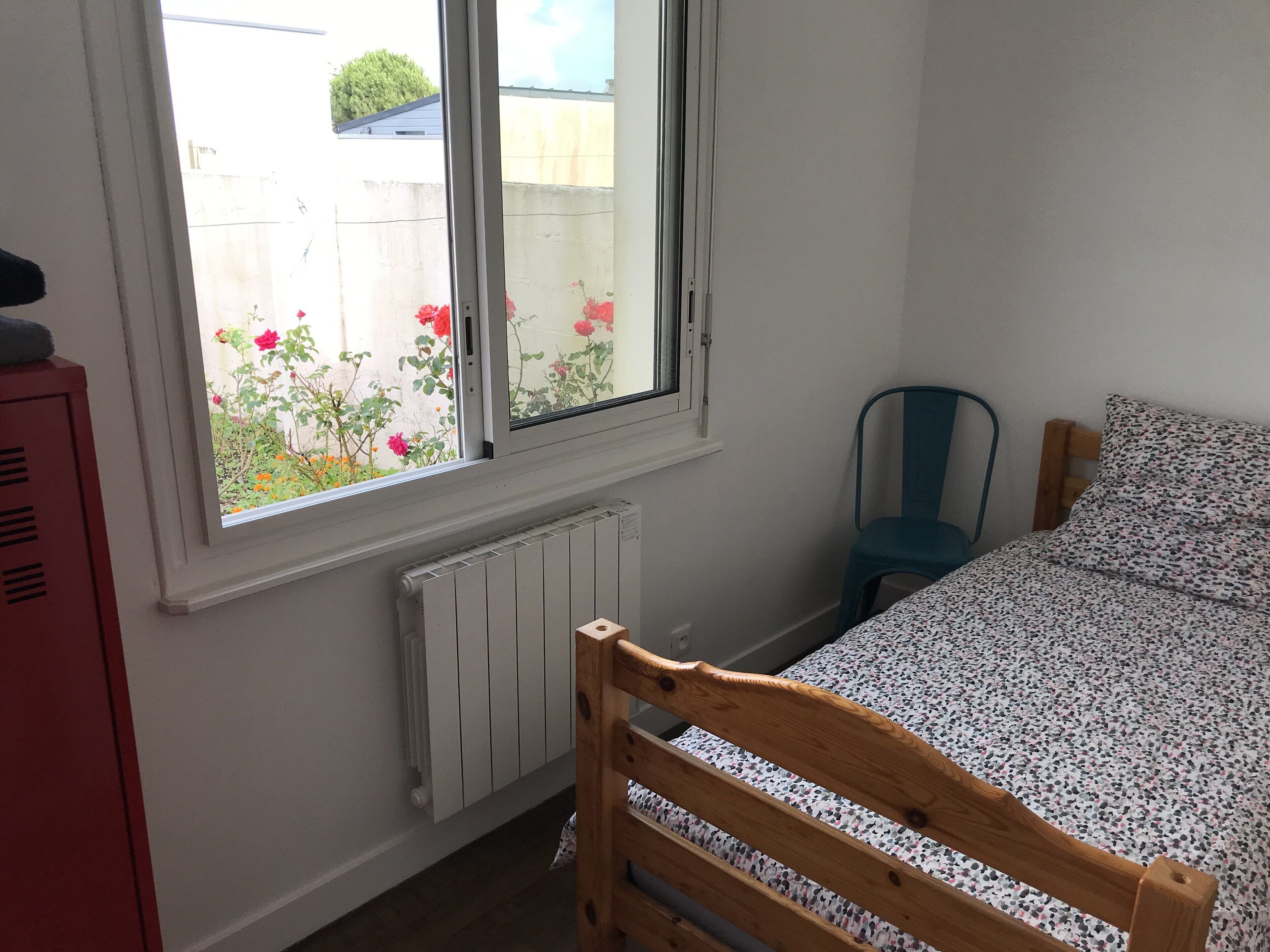 Une chambre avec un lit en bois, une fenêtre donnant sur un jardin, un radiateur et une chaise bleue.
