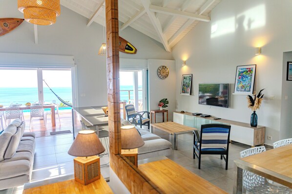 A spacious living area with a view of the ocean, a wooden dining table, and a TV mounted on the wall.