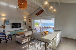 A modern kitchen with a large island, dining area with white chairs, and a view of the ocean through large windows.