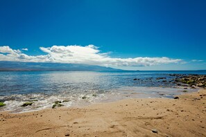 Ein Strand mit klarem, blauem Wasser, einem sandigen Ufer und Bergen im Hintergrund.