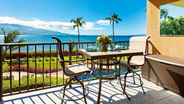 A balcony with a table, chairs, and a view of the ocean and mountains. - Endless Waves, Incredible Direct Oceanfront Views! (Wailuku)