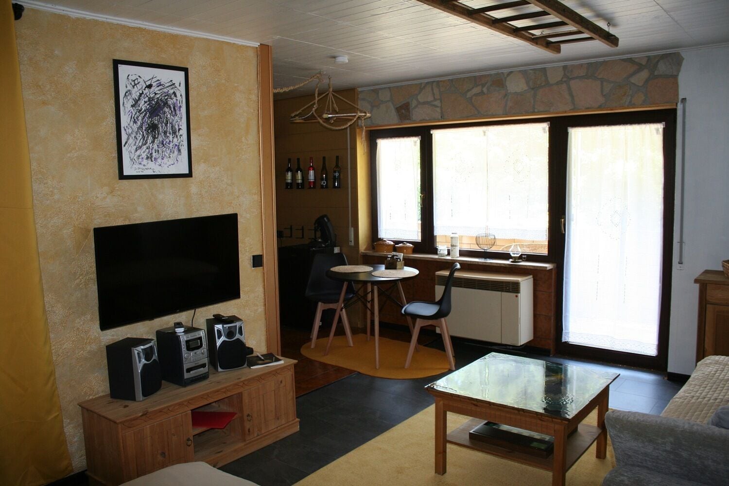 A living room with a flat-screen TV, a wooden entertainment unit, a dining area with a table and chairs, a kitchen area with a stove and window, and a stone wall.