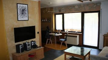 Un salon doté d’un téléviseur à écran plat, d’un meuble de rangement en bois, d’un coin repas avec une table et des chaises, d’une cuisine équipée d’une cuisinière et d’une fenêtre, et d’un mur en pierre.