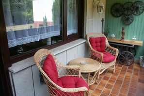Una silla de mimbre con un cojín rojo y una mesita redonda pequeña con un objeto de vidrio encima, ubicada en un patio con suelo de baldosas, pared verde y una ventana con cortinas de encaje.
