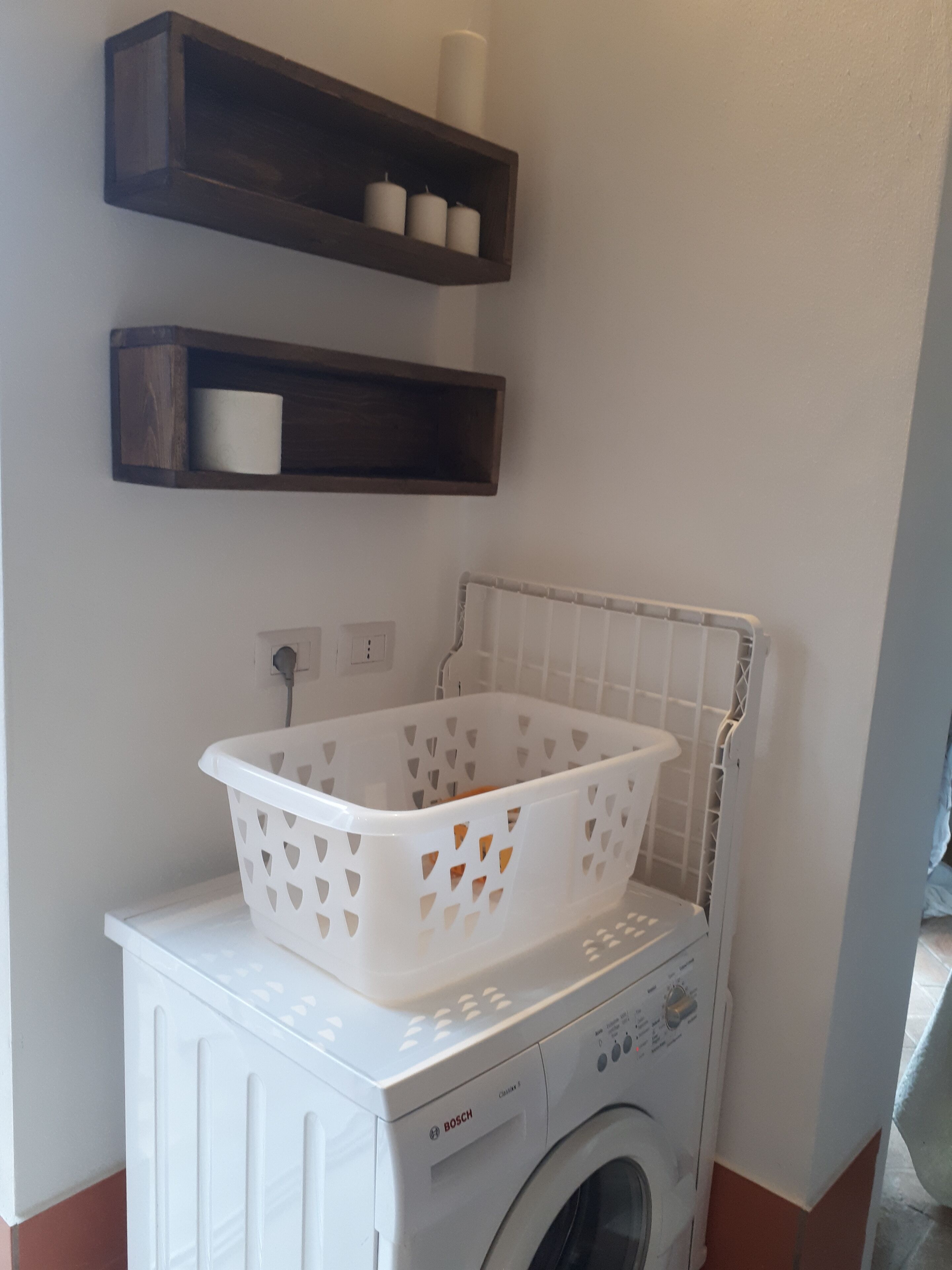 A laundry area with a white washing machine, a white laundry basket, and two dark wooden shelves with white candles.