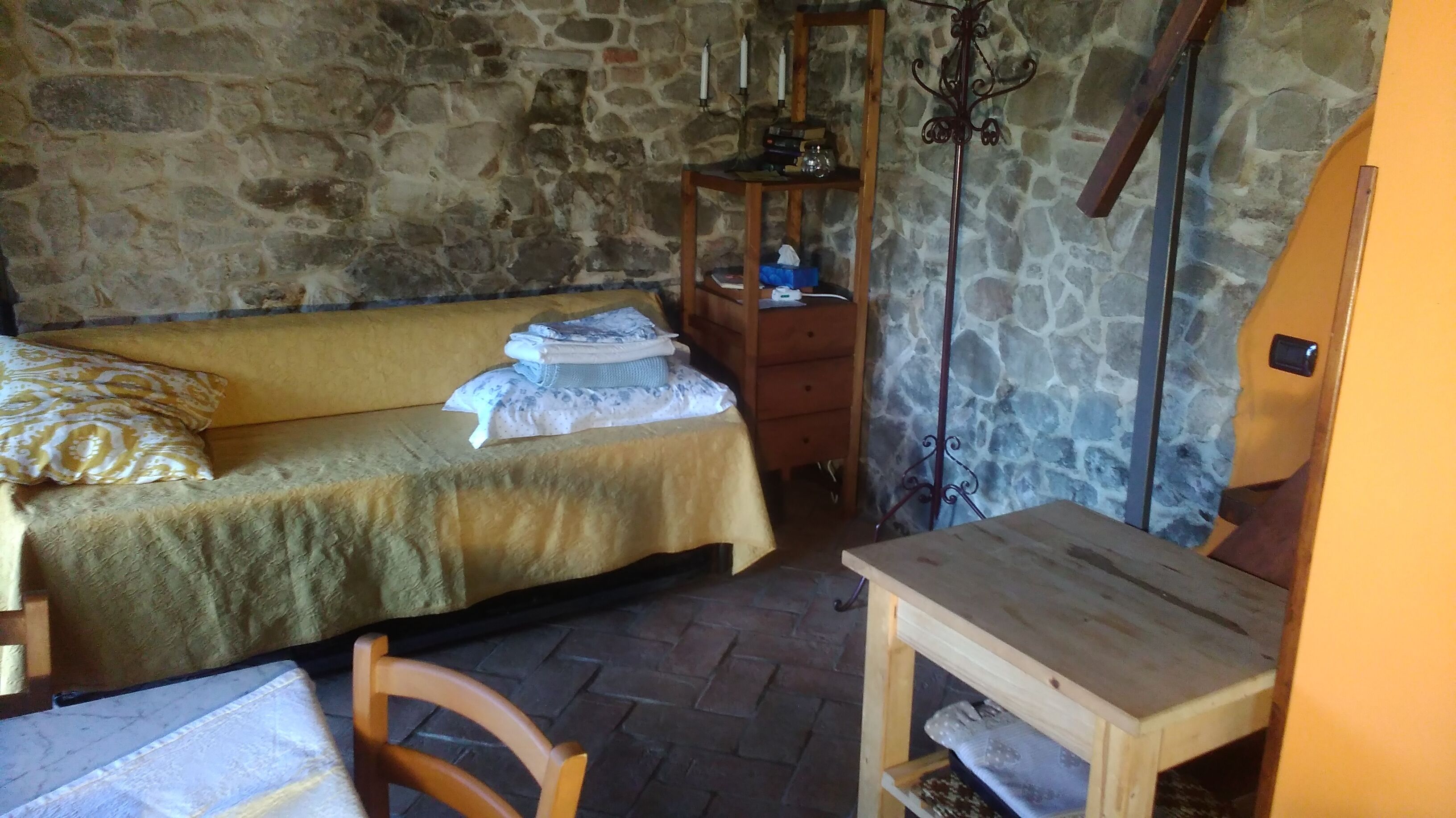 A small, cozy room with a stone wall, a wooden bed with a yellow bedspread, a wooden nightstand with a mirror, and a small wooden table.