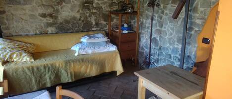 A small, cozy room with a stone wall, a wooden bed with a yellow bedspread, a wooden nightstand with a mirror, and a small wooden table.