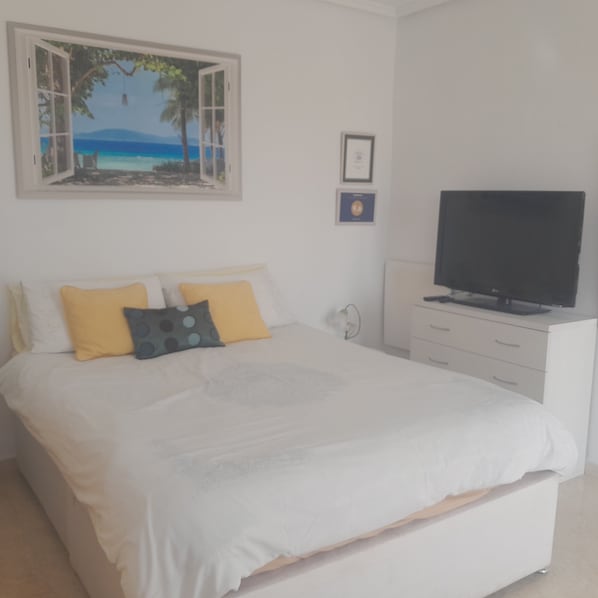 A bedroom with a bed, a TV on a stand, a painting of a beach, and a framed certificate on the wall.