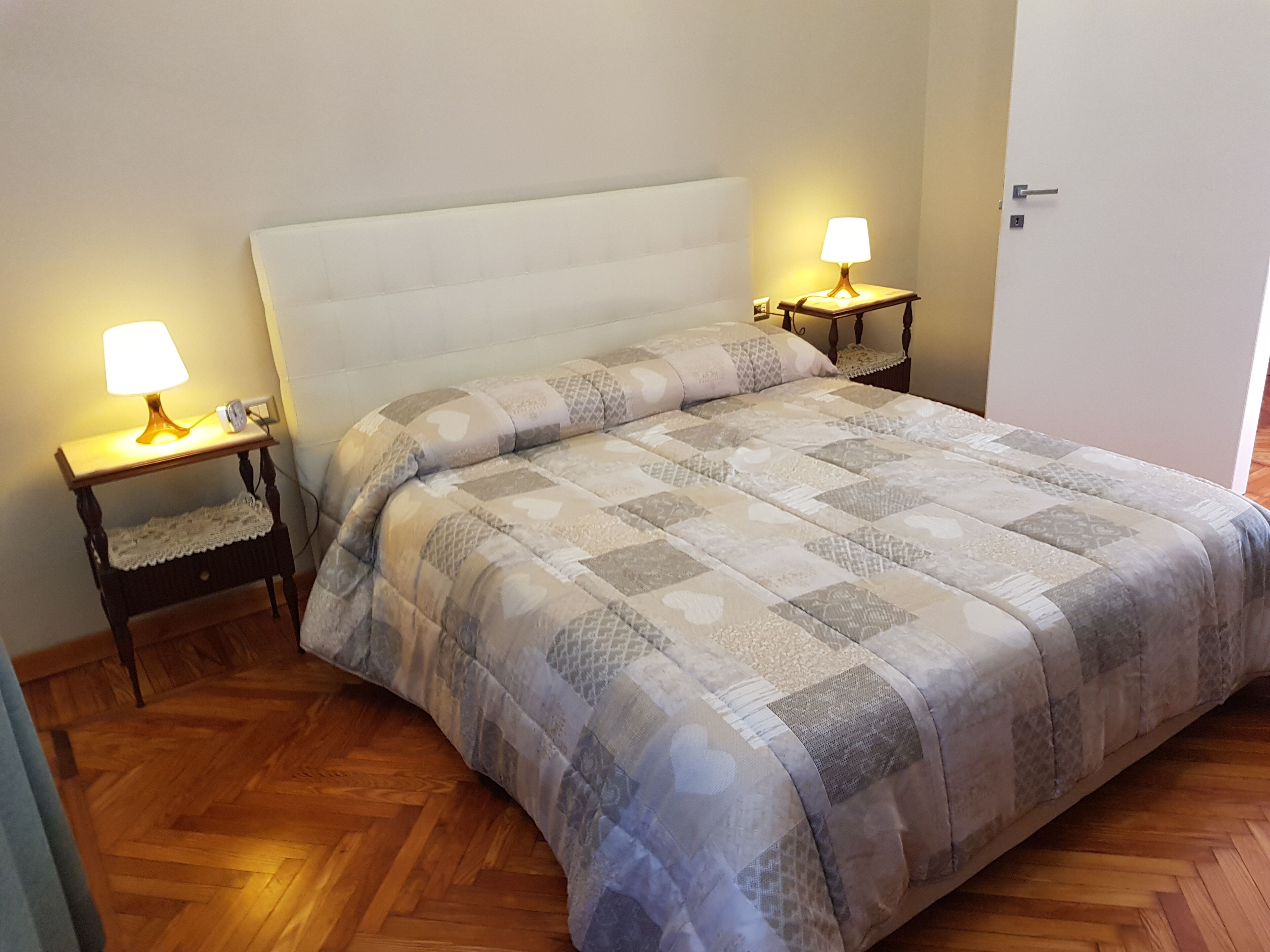 Un lit bien fait, avec une tête de lit matelassée, deux tables de chevet avec des lampes et un parquet en bois disposé en motif chevrons.