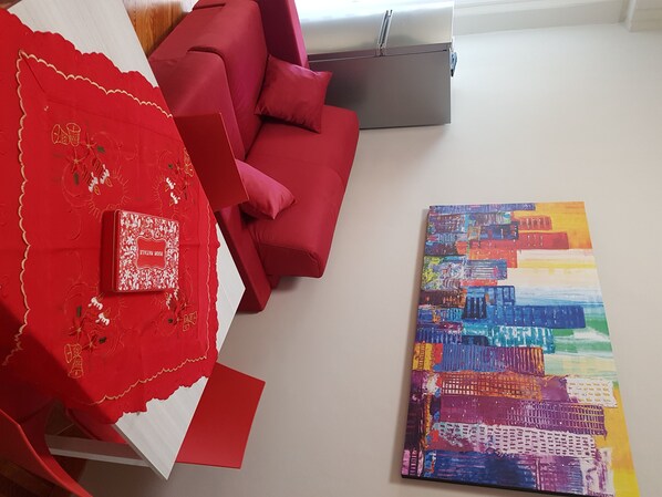 A room with a red sofa, a colorful abstract painting, and a red patterned tablecloth.