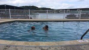 A swimming pool with two people in the water, surrounded by a white fence and a parking area in the background.