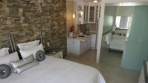Un dormitorio con una cama grande, una mesita de noche, un baño visible a través de una puerta abierta y una zona de cocina.