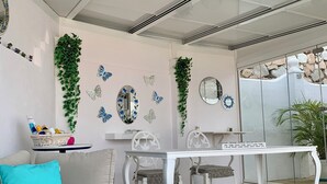 Una habitación con una mesa blanca, dos sillas, un sofá con un cojín azul, mariposas decorativas y plantas en las paredes.
