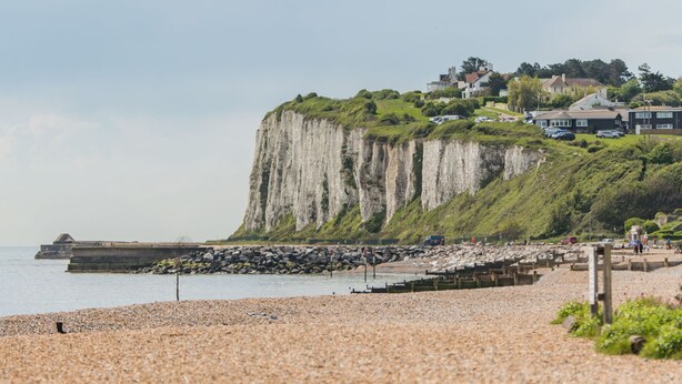 Ein Kiesstrand mit einer Klippe und Häusern im Hintergrund.