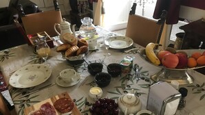 Una mesa de comedor servida con diversos alimentos, como pan, fruta, queso y embutidos, además de tazas de té y café.